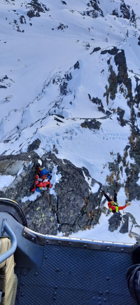 Zo záchrannej akcie HZS - záchranár HZS v podvese vrtuľníka nad pacientom s ďalším záchranárom HZS na zasneženom skalnatom teréne 