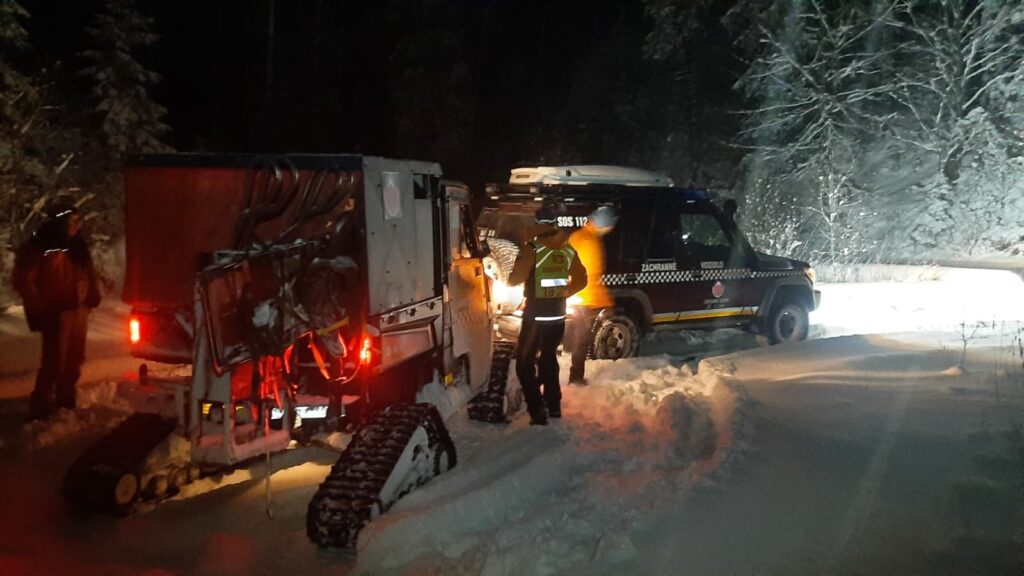terénne vozidlo HZS na pásoch na zasneženom TZCH v noci, pri vozidle stoja záchranári, pred pásovým vozidlom stojí ďalšie terénne záchranné vozidlo HZS 