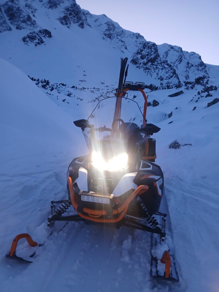 snežný skúter HZS v zasneženom teréne, v pozadí zasnežený skalný hrebeň Západných Tatier 