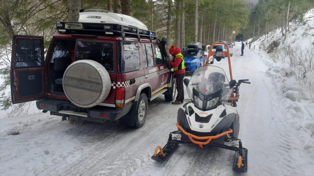 terénne vozidlo HZS na zasneženej lesnej ceste, pri vozidle stojí záchranár HZS a  vedľa vozidla odstavený snežný skúter HZS 