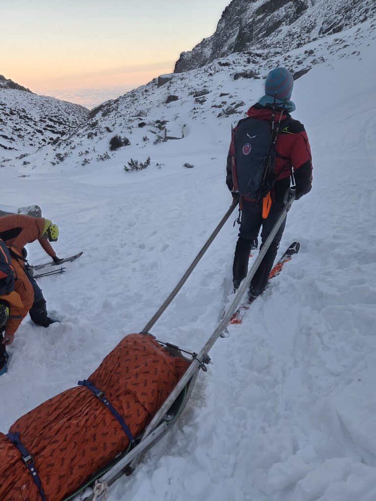 horskí záchranári transportujú pacienta - pacient na saniach, zateplený, záchranári na lyžiach 