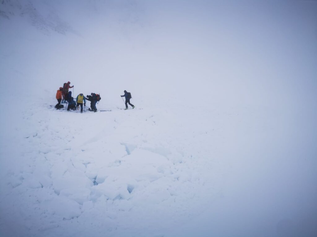 skupina svedkov na spodku lavínišťa pri zranenom turistovi, poskytnutie prvej pomoci a zateplenie, v pozadí zasnežené skalné masívy a hmla 