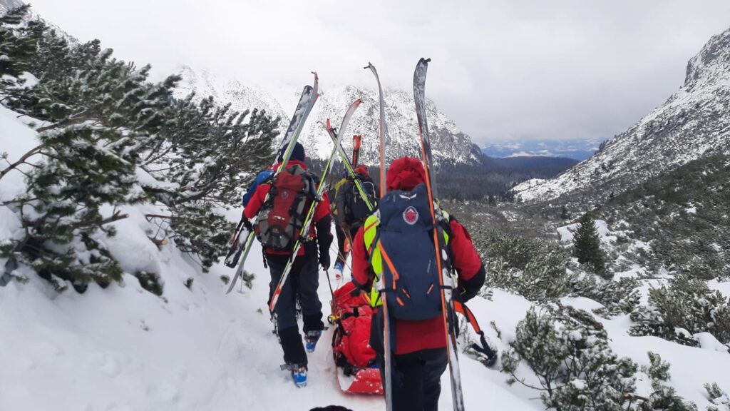 záchranári HZS transportujú pacienta pozemne zasneženou dolinou, lyže pripnuté na batohoch 