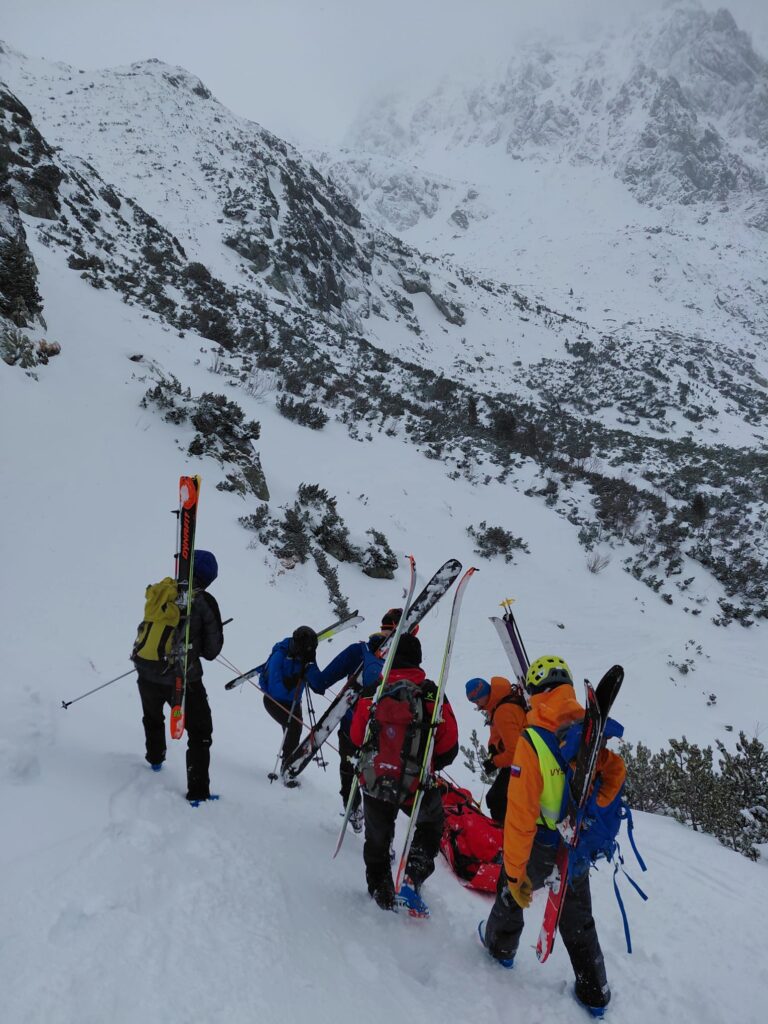 transport pacienta pomerne strmou časťou zasneženej doliny , pacient nabalený v transportnom prostriedku, zaistený lanovou technikou,