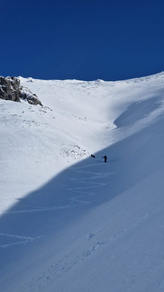 záchranár HZS so psom smerujúci hore žľabom na lyžiach (lyžami vytvorené cik-caky), v pozadí modré nebo