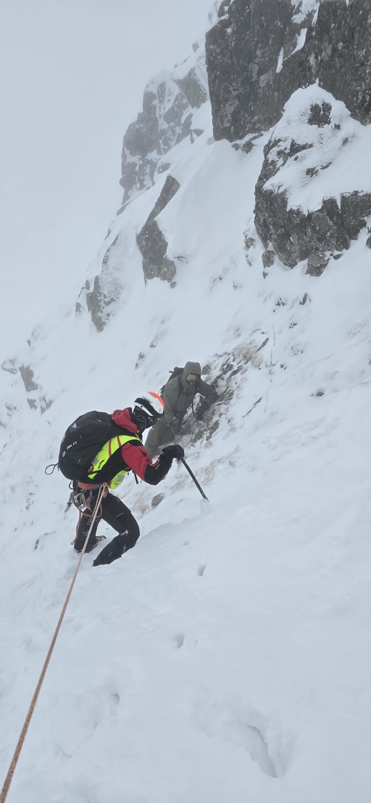 záchranár HZS v exponovanom skalnom zasneženom teréne na lane, s cepínom, prichádza k uviaznutému mužovi