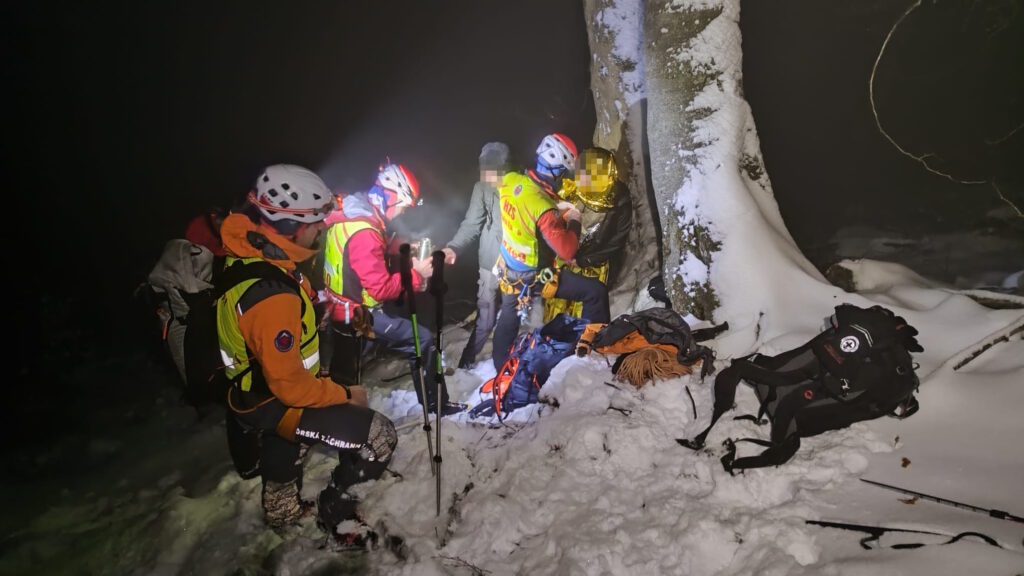 horskí záchranári zatepľujú zablúdených turistov, tma a sneh, záchranári si svietia čelovkami, jedného z turistov akurát balia do termofólie 