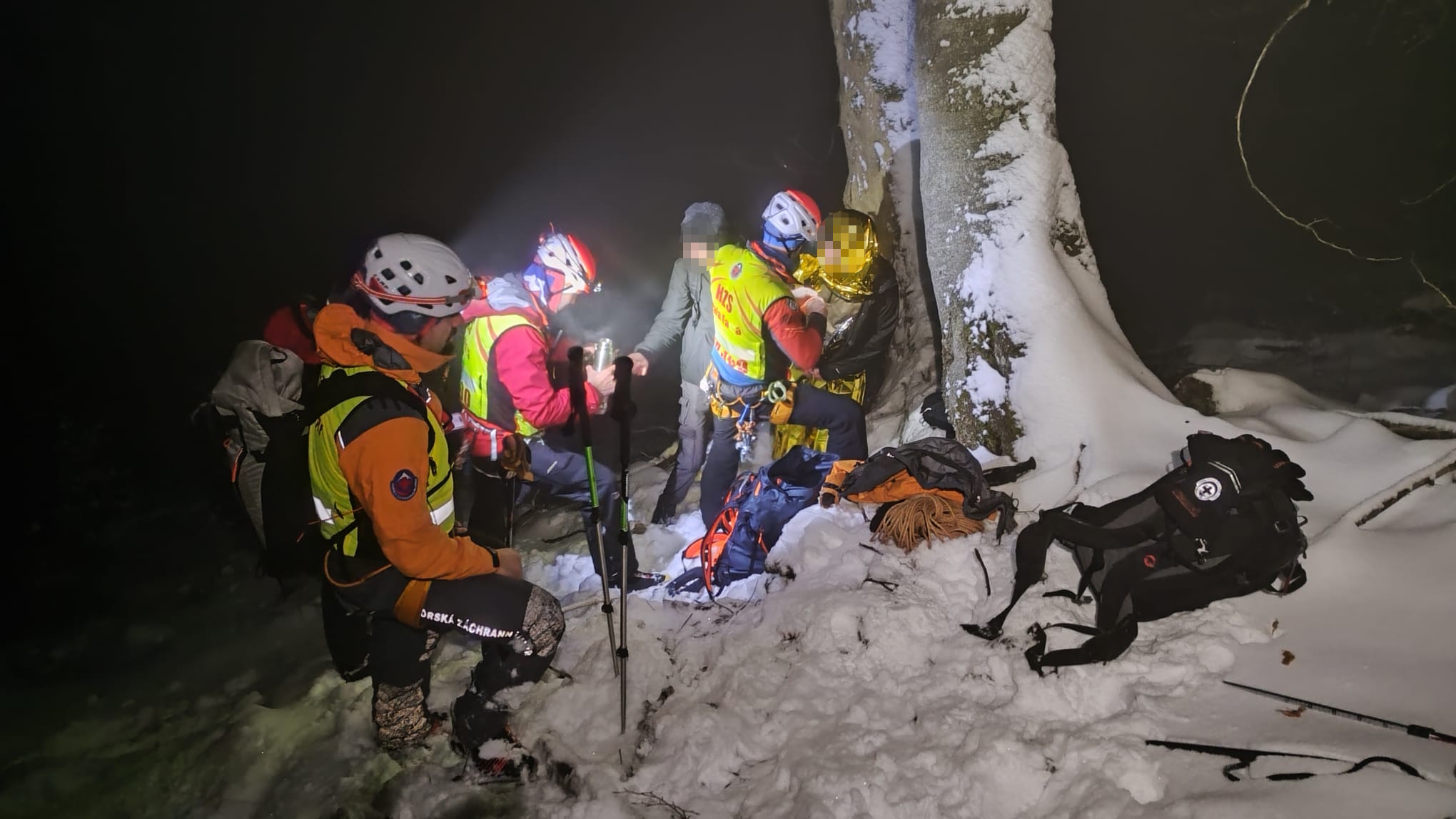 horskí záchranári zatepľujú zablúdených turistov, tma a sneh, záchranári si svietia čelovkami, jedného z turistov akurát balia do termofólie
