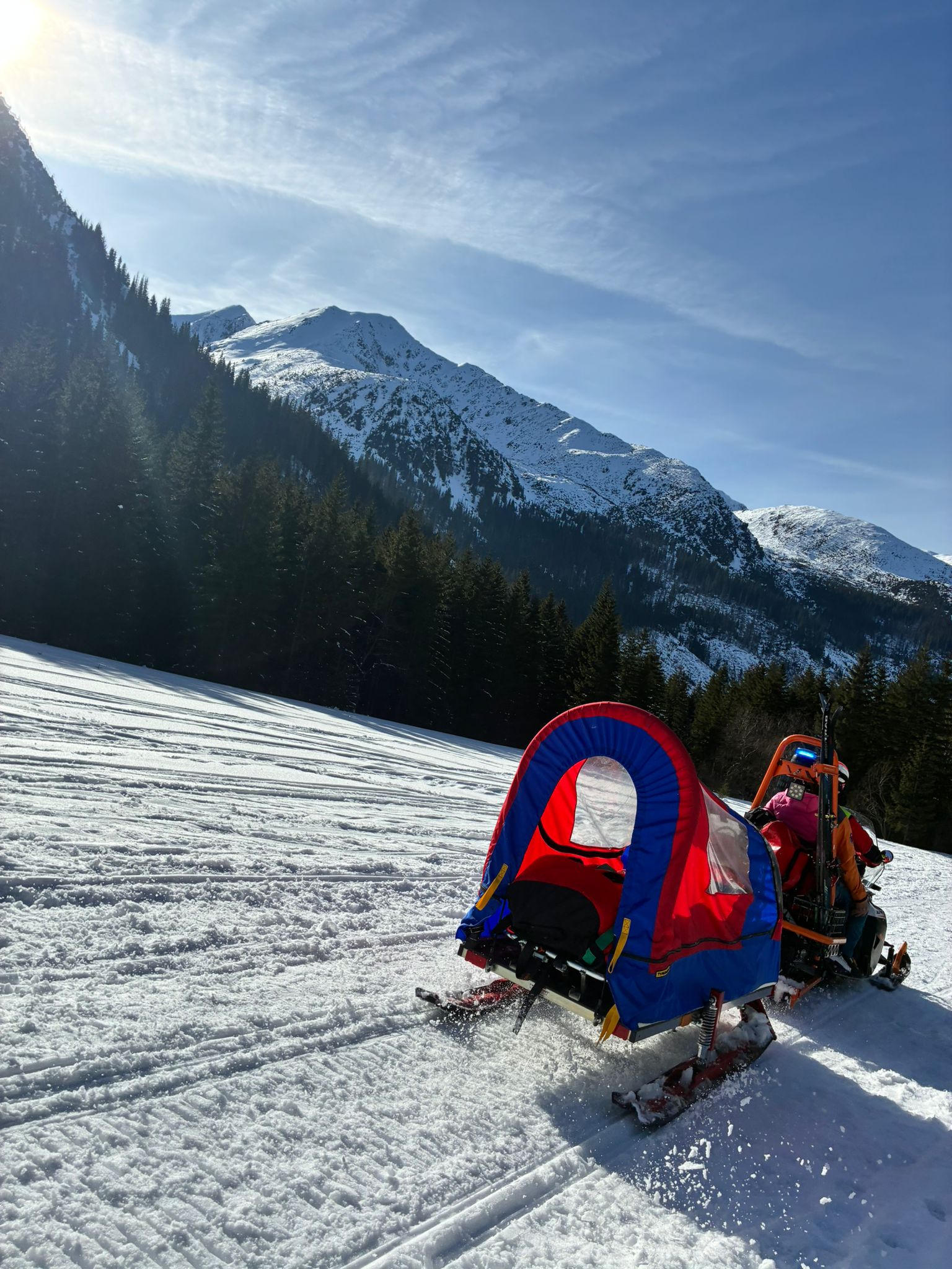 záchranár HZS na skútri v zasneženej doline, za skútrom sú pripnuté sane s pacientom, v pozadí vrcholy Západných Tatier