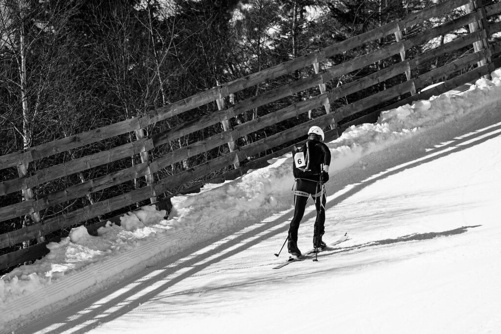 čiernobiela fotografia zachytávajúca výstup pretekára na skialpinistických pásoch popri drevených zábranách na zjazdovke 