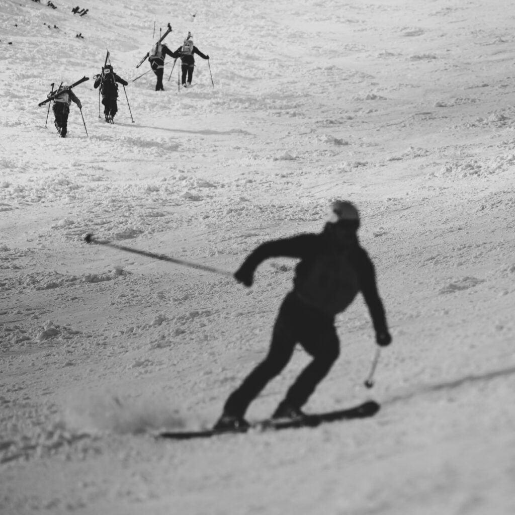 čiernobiela fotografia zachytávajúca výstup pretekárov na mačkách s lyžami na chrbte smerom do Lomnického sedla, jeden pretekár už lyžuje smerom do cieľa 