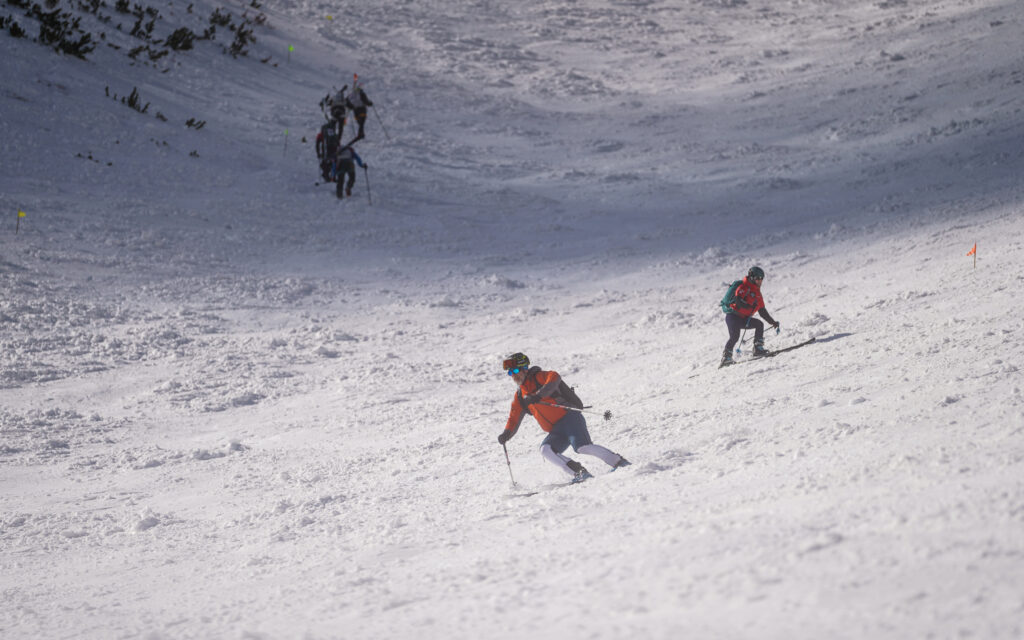 jedna dvojica pretekárov lyžuje smerom do cieľa, ďalší pretekári vystupujú s lyžami na chrbte