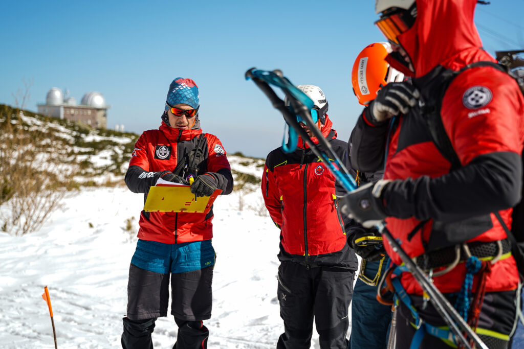 pretekár vyhľadáva lavínovým vyhľadávacím prístrojom, jeden organizátor zapisuje čas, druhý organizátor kontroluje správnosť postupu 