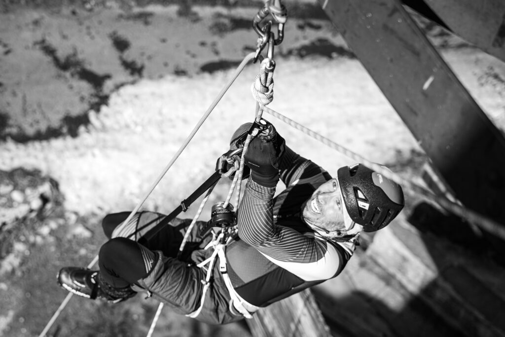 zhora odfotený pretekár naviazaný na lane vystupujúci po lane pomocou žumaru, čiernobiela fotografia, pretekár je už úplne na vrchu 