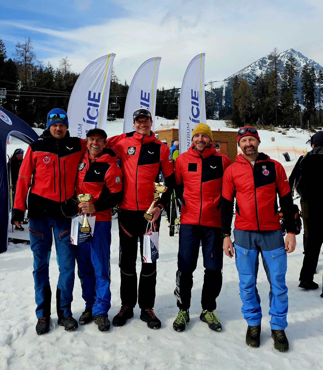 na fotke sú šiesti záchranári HZS, ktorí sa zúčastnili preteku, v červených služobných bundách, v rukách dvaja z nich majú trofeje za prvé miesto, v pozadí areál Štrbského plesa, zľava Erik Rabatik, Ján Šustr, Štefan Hudák, Peter Diča, Martin Matúšek
