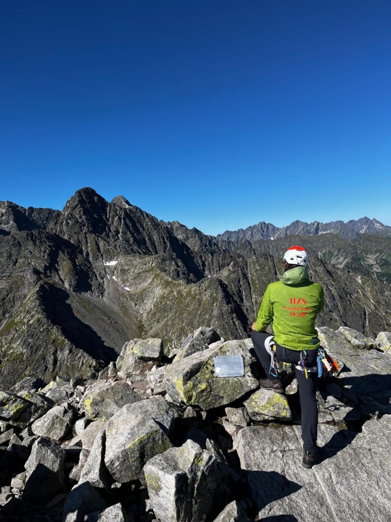 ilustračná foto - záchranár HZS v zelenej bunde na vrchole tatranského štítu, v pozadí modré nebo a skalné masívy