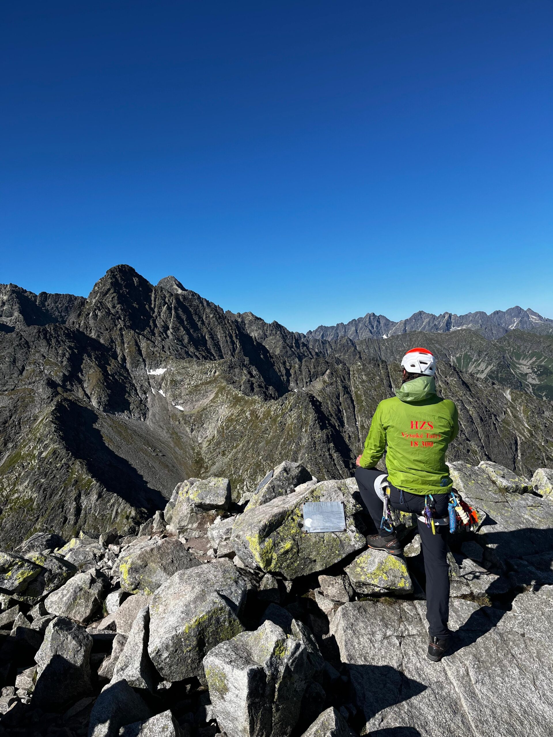 ilustračná foto - záchranár HZS v zelenej bunde na vrchole tatranského štítu, v pozadí modré nebo a skalné masívy