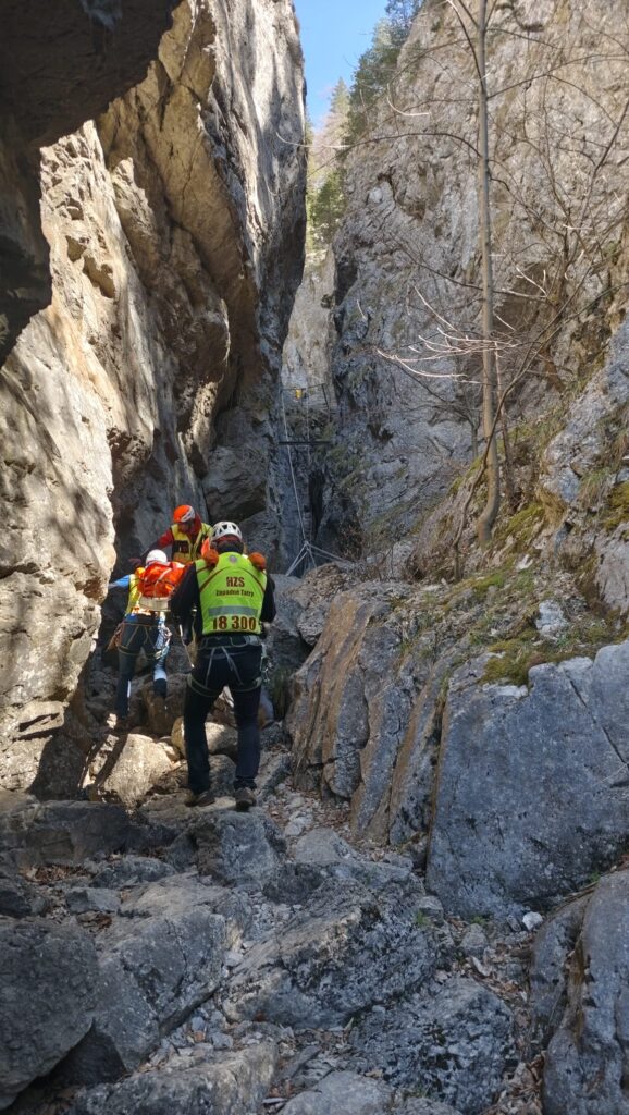 záchranári HZS smerujúci hore dolinou po exponovanom teréne s pacientom na nosidlách 