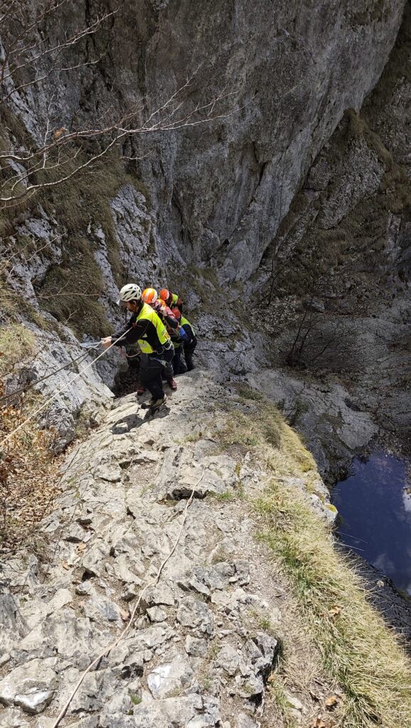 záchranári HZS v exponovanom teréne s pacientom na nosidlách stúpajúci po skalnom teréne zabezpečenom oceľovými lanami 