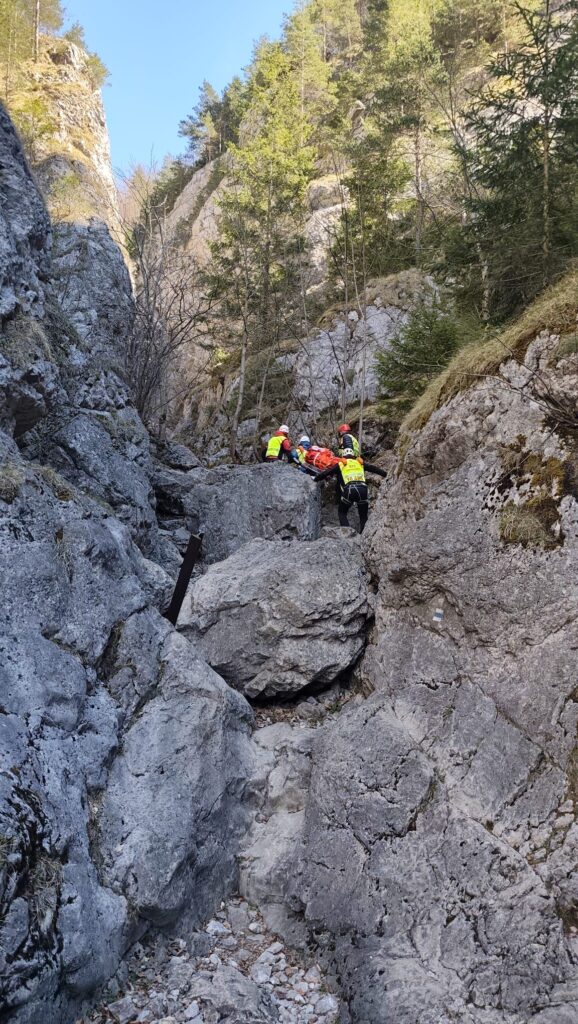 záchranári HZS smerujúci hore dolinou po exponovanom teréne s pacientom na nosidlách, v okolí skalné bralá 
