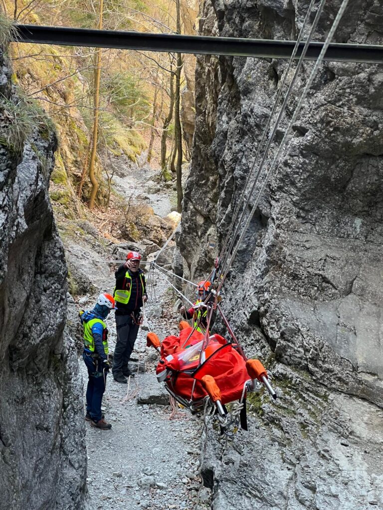 pacient na nosidlách v Prosieckej doline, nosidlá sú vo vise a spúšťané pomocou lanovky do terénu. kde je možný klasický transport, pri lanovke stoja traja záchranári 